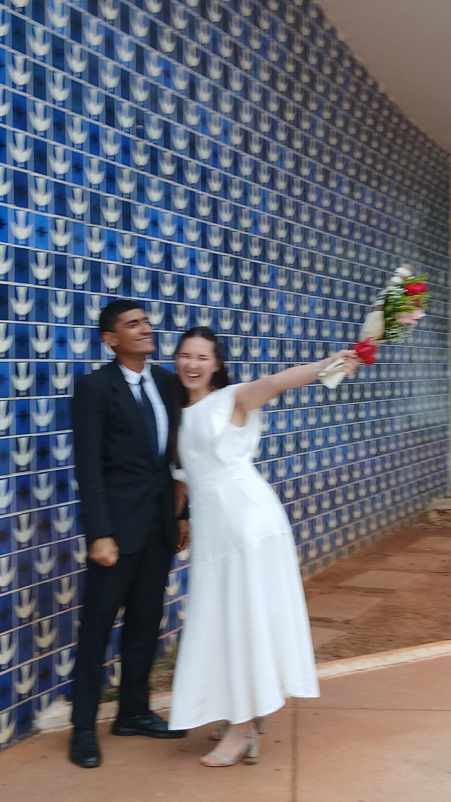 Aline e Arthur sorrindo com o buquê diante de um painel de azulejos azuis